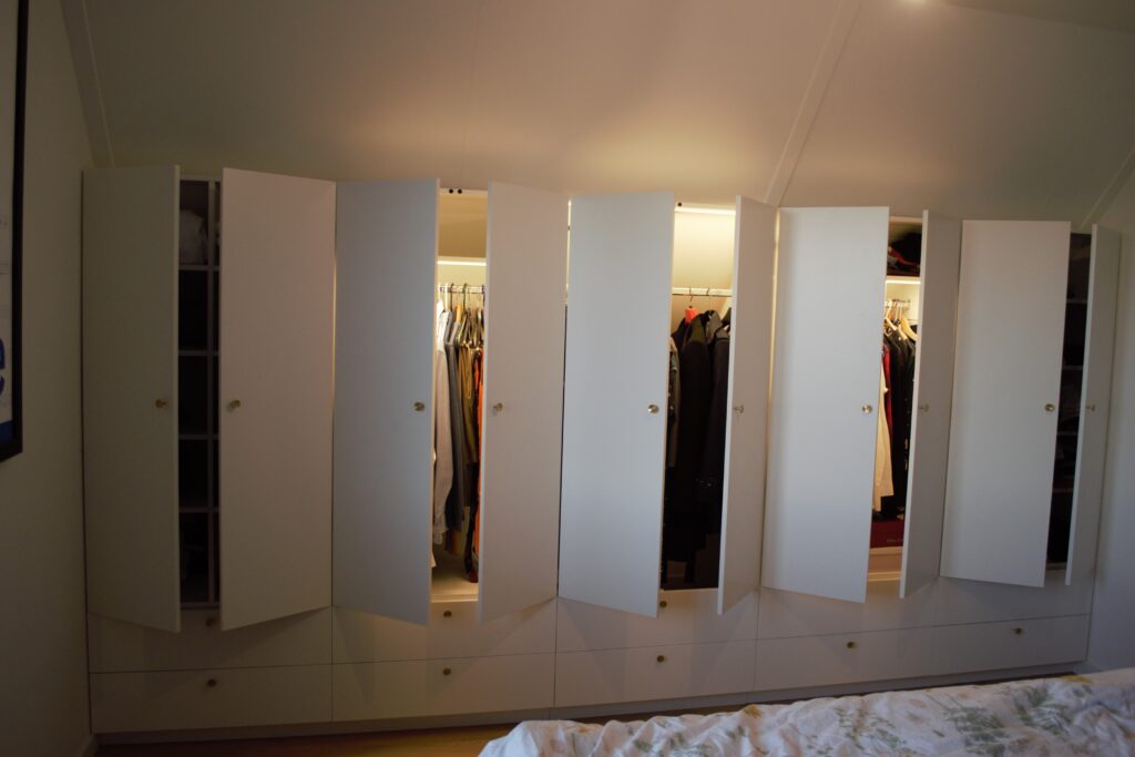 Kastenwand over de gehele breedte van een master bedroom, met geïntegreerde LED-verlichting en subtiele, ronde knoppen van massief geborsteld messing. Het middendeel van de kast, inclusief de laden, is uitneembaar en biedt toegang tot opbergruimte onder het dakschot! #kastopmaat #maatwerk #interieurbouw #abcoude #landvanwinkel #wardrobe #interior #interiordesign #masterbedroom #residential #residentialarchitecture #bespoke #bespokewardrobe #bespokefurniture #custommade @abcoude.nl